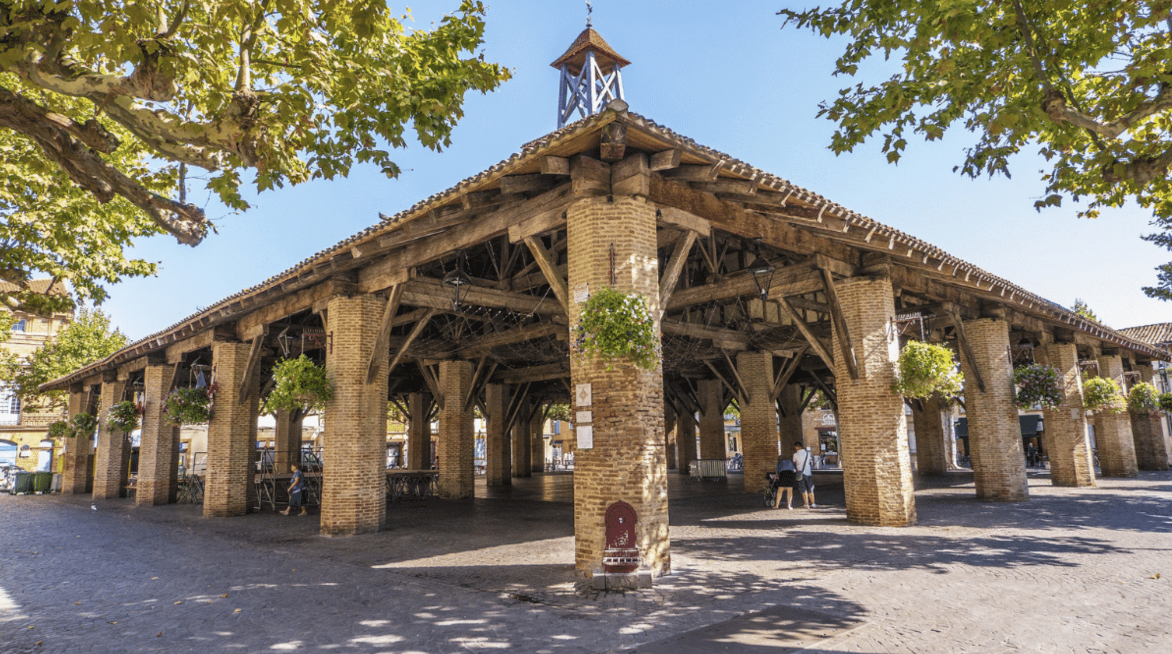 Halle médiévale de Grenade avec piliers en briques et charpente bois sous un ciel bleu ensoleillé en Haute-Garonne.