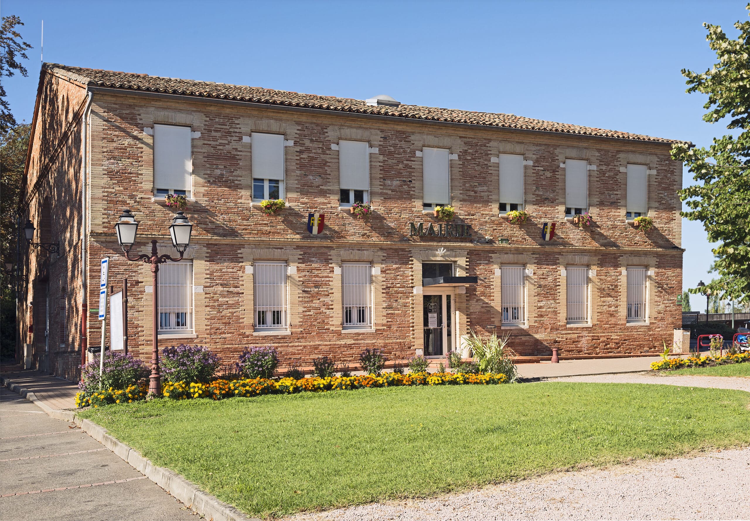 Mairie en briques rouges avec emblèmes RF, volets blancs et parterre de fleurs sous un ciel bleu.