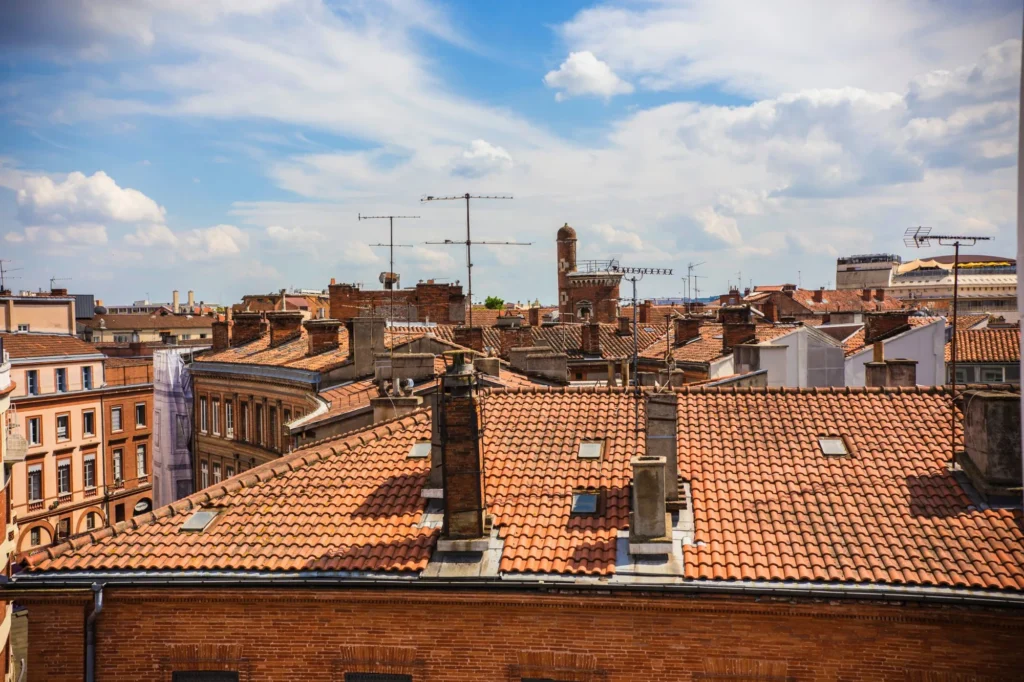 Toits de Toulouse : Briques Roses et Tuiles Rouges Toits en tuiles rouges et façades en briques de Toulouse sous un ciel bleu parsemé de nuages blancs.