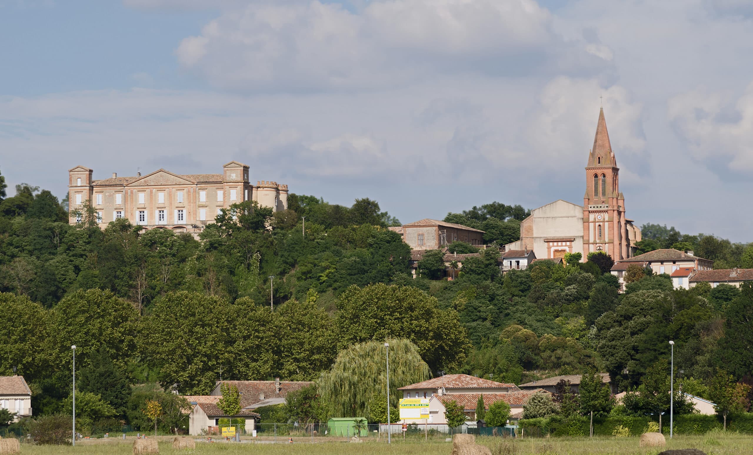 Château et église en briques roses dominant un village d'Occitanie (Castelnau-d'Estrétefonds) entouré de végétation sous un ciel bleu nuageux.