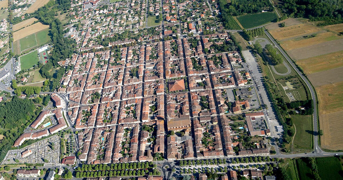 Vue aérienne d'une bastide du Sud-Ouest au plan en damier, toits de tuiles rouges et champs environnants.