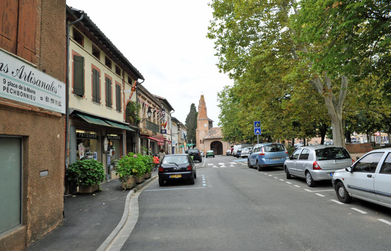 Pechbonnieu : Rue commerçante typique en Haute-Garonne Rue de Pechbonnieu avec boulangerie-pâtisserie, voitures stationnées et clocher de l'église au bout de la chaussée.